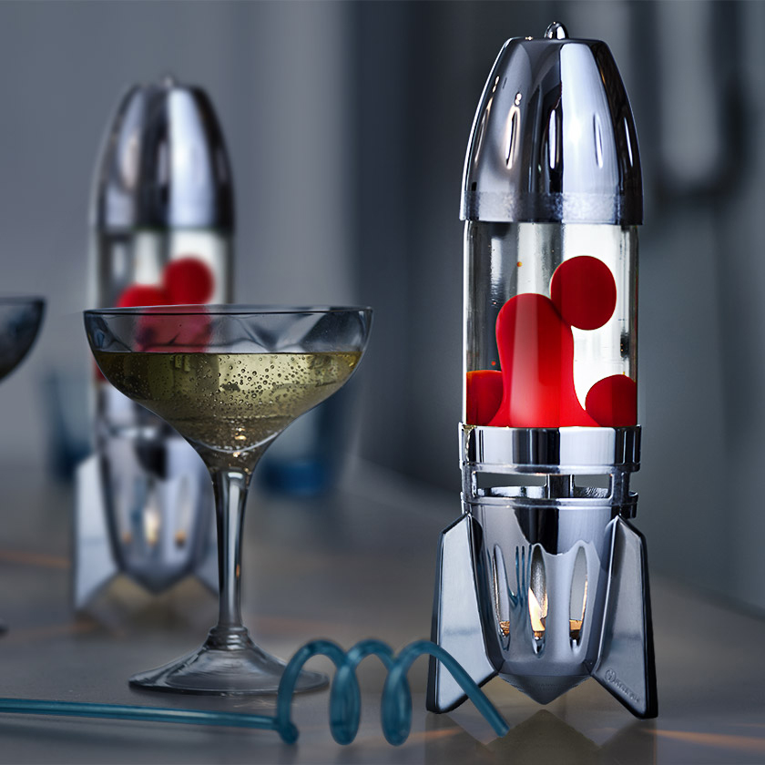Sidabrinė raketos formos Mathmos Fireflow žvakinė lavos lempa su skaidriu skysčiu ir raudona lava ant stalo šalia kokteilio taurės