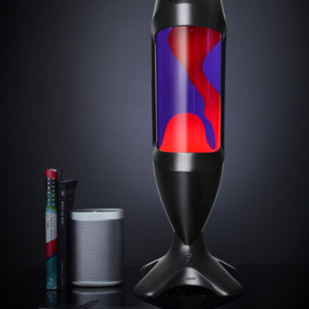 Originali Mathmos iO didelė lavos lempa – Juoda / Violetinė / Raudona Didelė juoda Mathmos iO lavos lempa su violetiniu skysčiu ir raudona lava tamsiame fone šalia knygų ir garsiakalbio